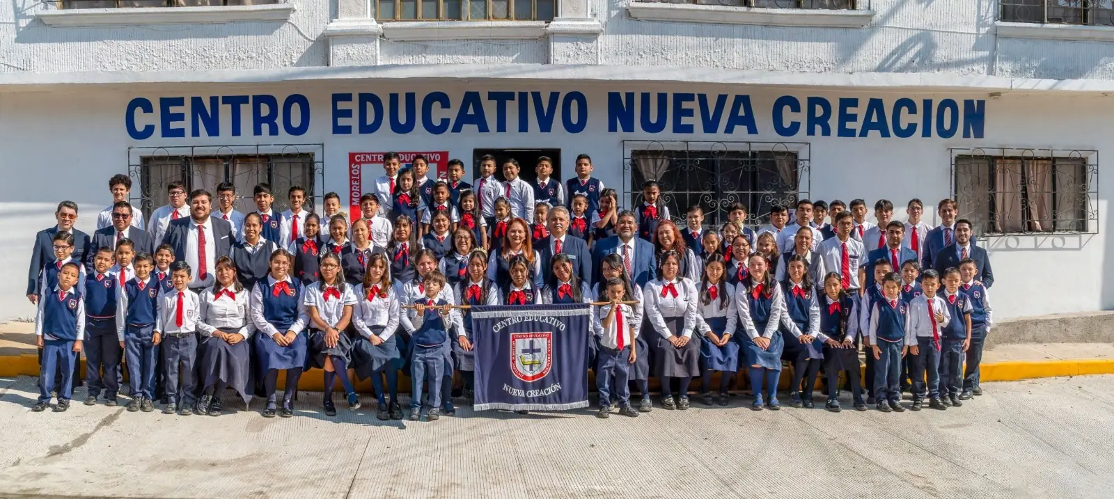 Alumnos, maestras, maestros y directivos de CENCREA con uniforme frente a la fachada de la escuela, con el letrero «Centro Educativo Nueva Creación» y el estandarte del colegio